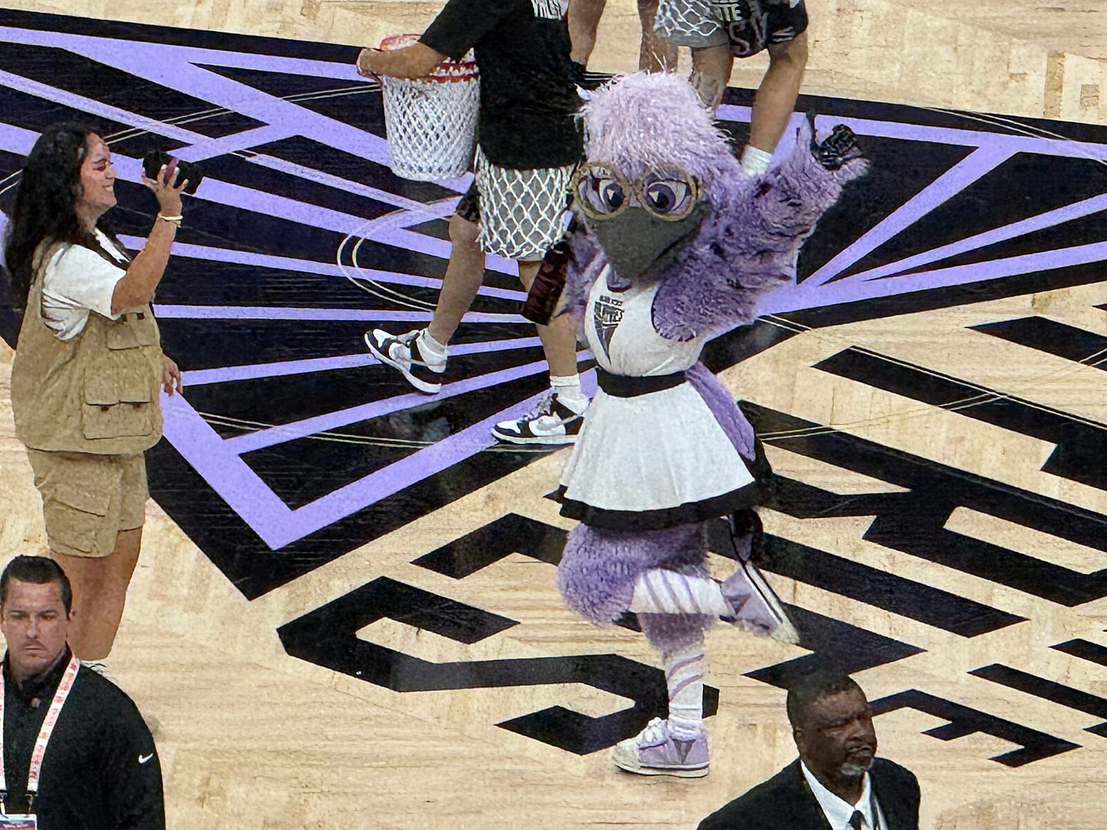 A purple feathered bird mascot in a little white skirt strikes a pose. Around her are bouncers and one person wearing a khaki zookeeper outfit.