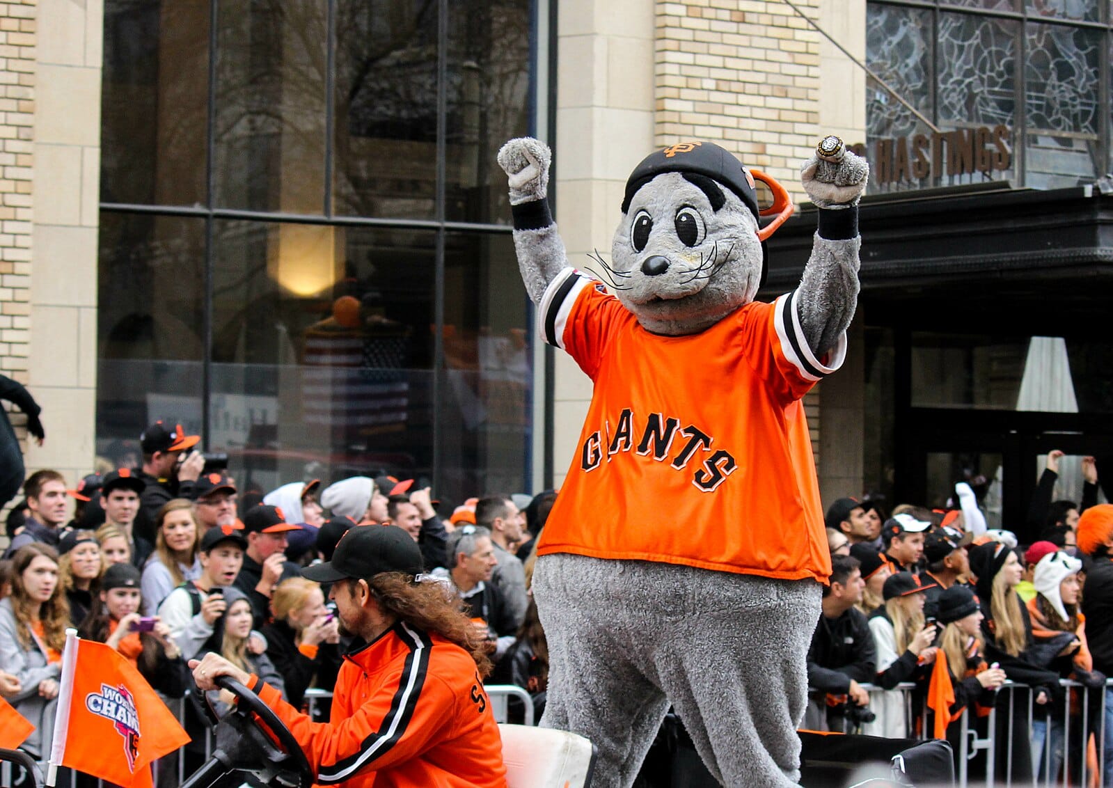 A person in a grey furry costume and an orange giants jersey holding up their arms while riding on the back of a golf cart.