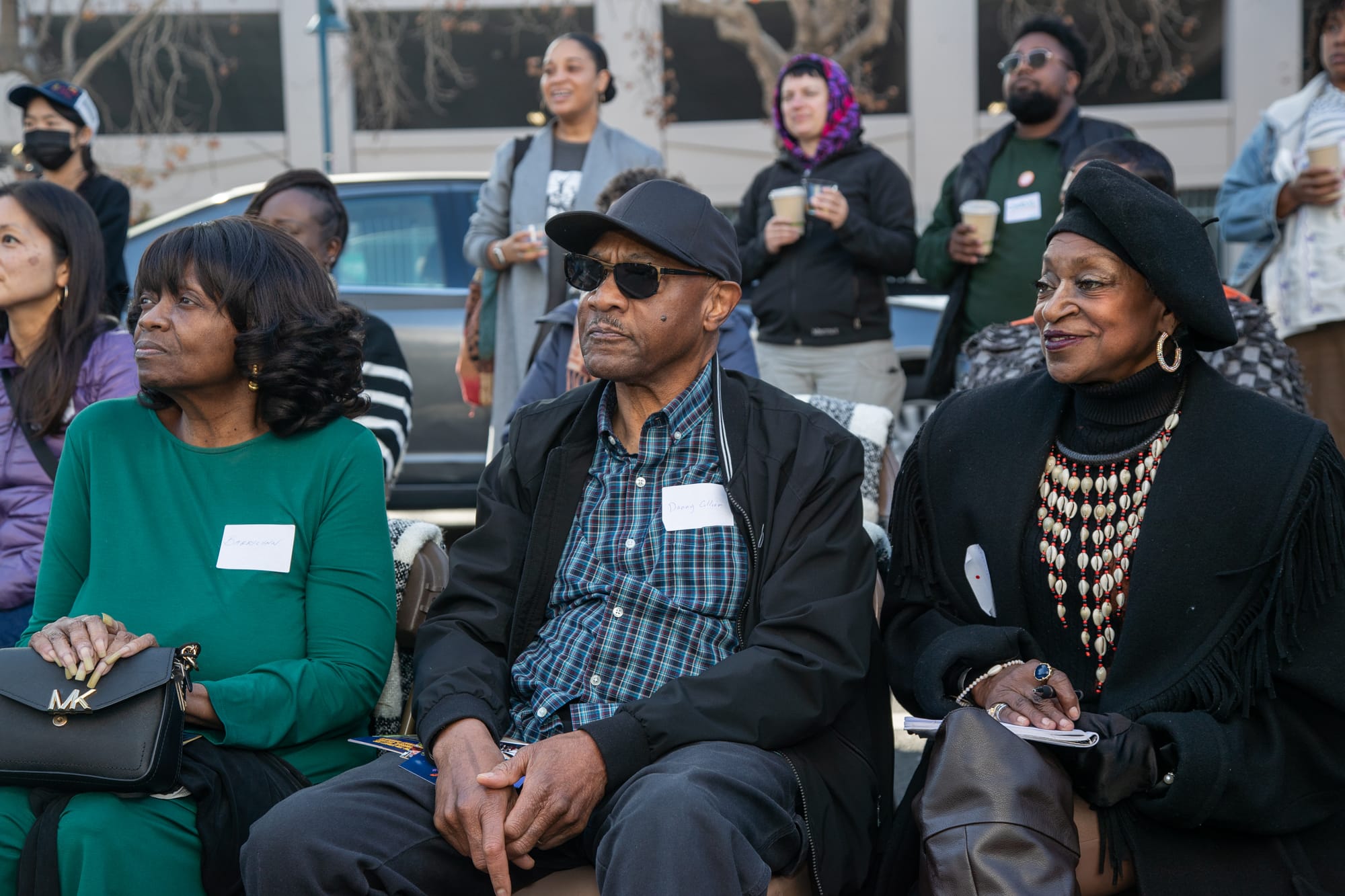 Three Black people sit in chairs looking off to the left (at the stage, off screen). In the middle, the man is wearing a black baseball hat, sunglasses, and a blue and black checked shirt.