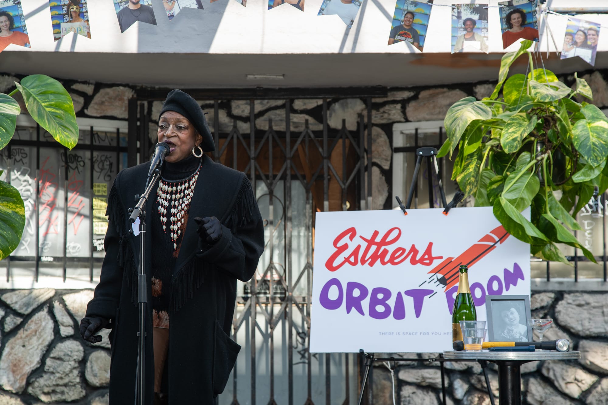 A Black woman in a black coat, beret, gold hoops and a dramatic seashell necklace sings into a microphone. In the background is a sign for Esthers Orbit Room.