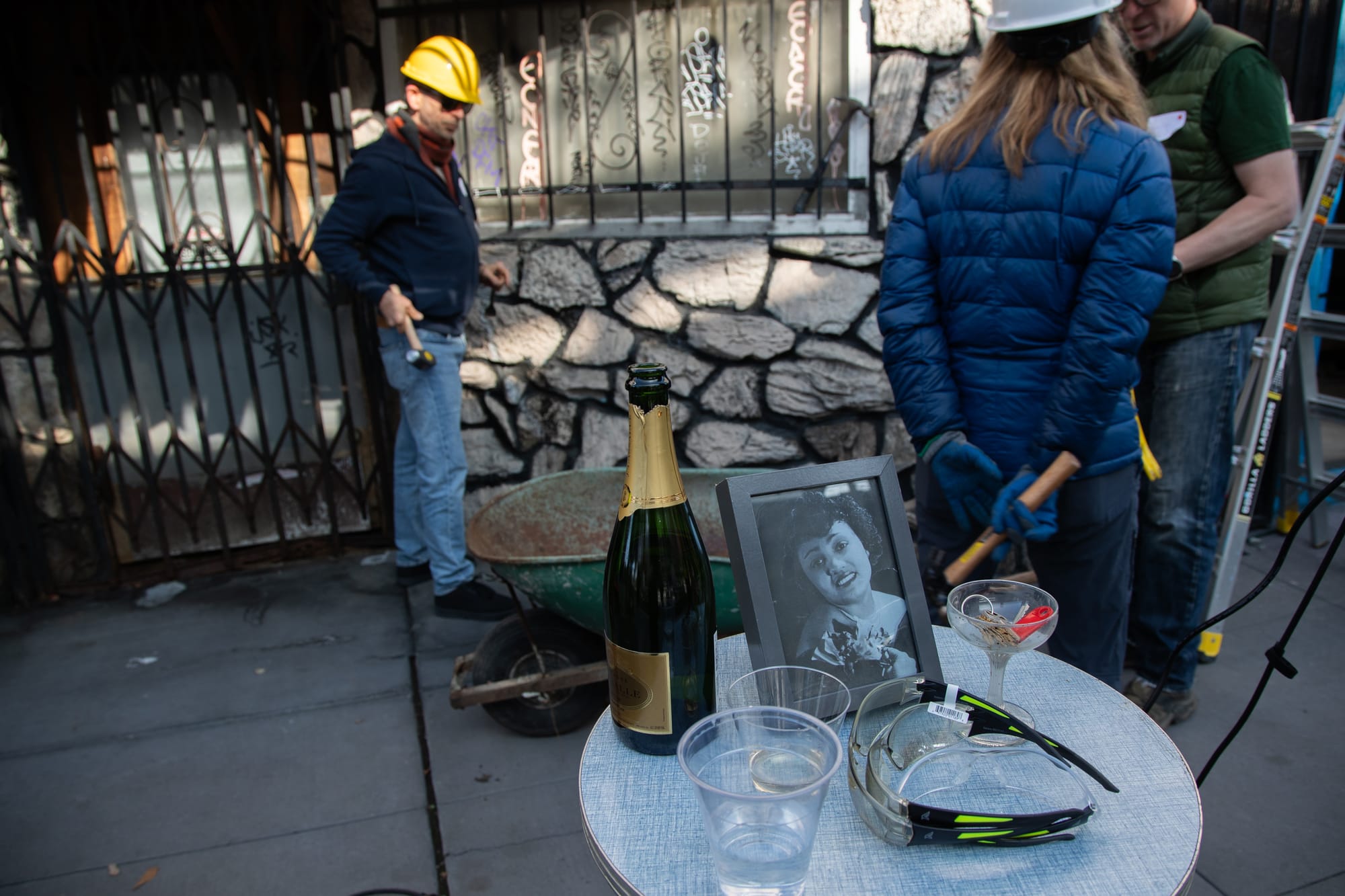 In the foreground, a small round table that has a campaign bottle, several safety glasses, a back and white photograph of Esther Mabry, and a small coup glass with keys inside on it. In the background, several people stand with hammers and hardhats, around a wheelbarrow. 