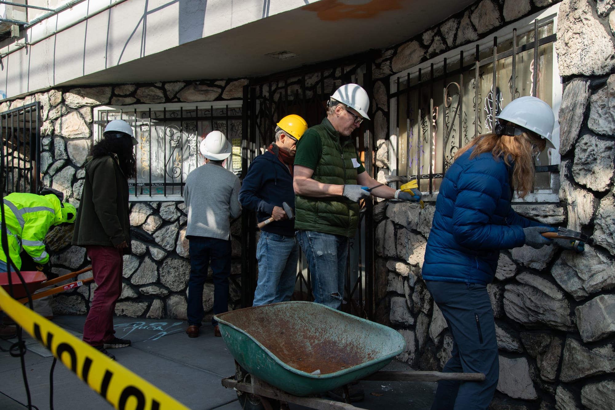 Several people stand in front of a stone wall, using hammers and chisels to try to get the stones out. In the center is a white man wearing a green puffy vest and a white hardhat.