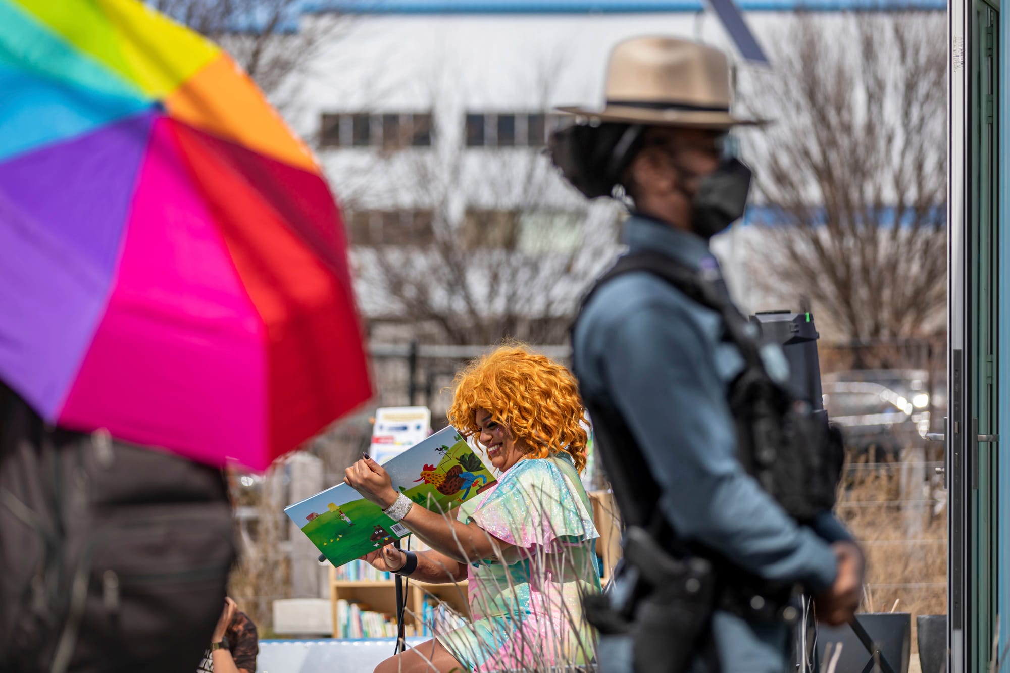 a police officer wearing a bulletproof vest is in the foreground while a drag queen smiles and reads a children's book in the background at an outdoor event