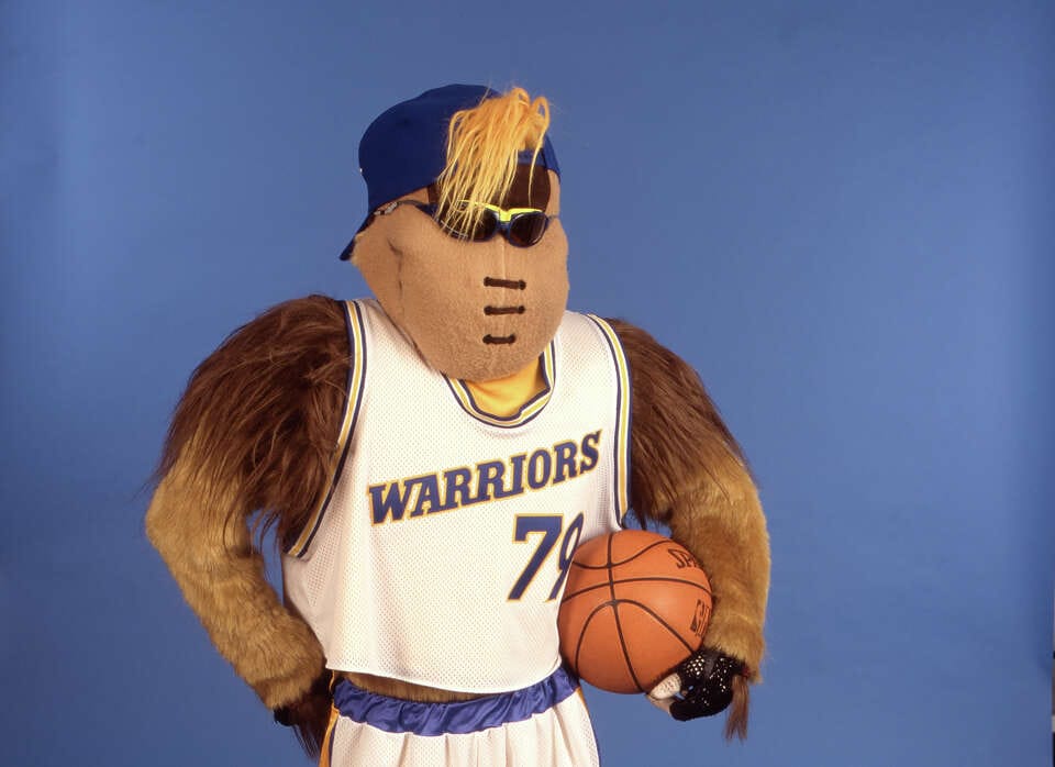 A mascot in front of a blue background holding a basketball, wearing a Warriors jersey. It's a kind of bird type animal?