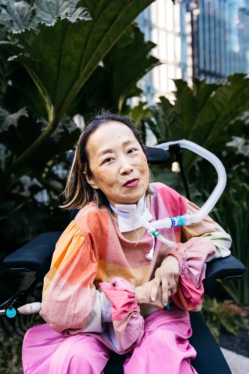 Alice Wong, Asian American woman in a wheelchair with a tracheostomy at her neck connected to a ventilator. She’s wearing a pink plaid shirt, pink pants, and a magenta lip color. She is smiling and behind her are a bunch of tall prehistoric looking plants.