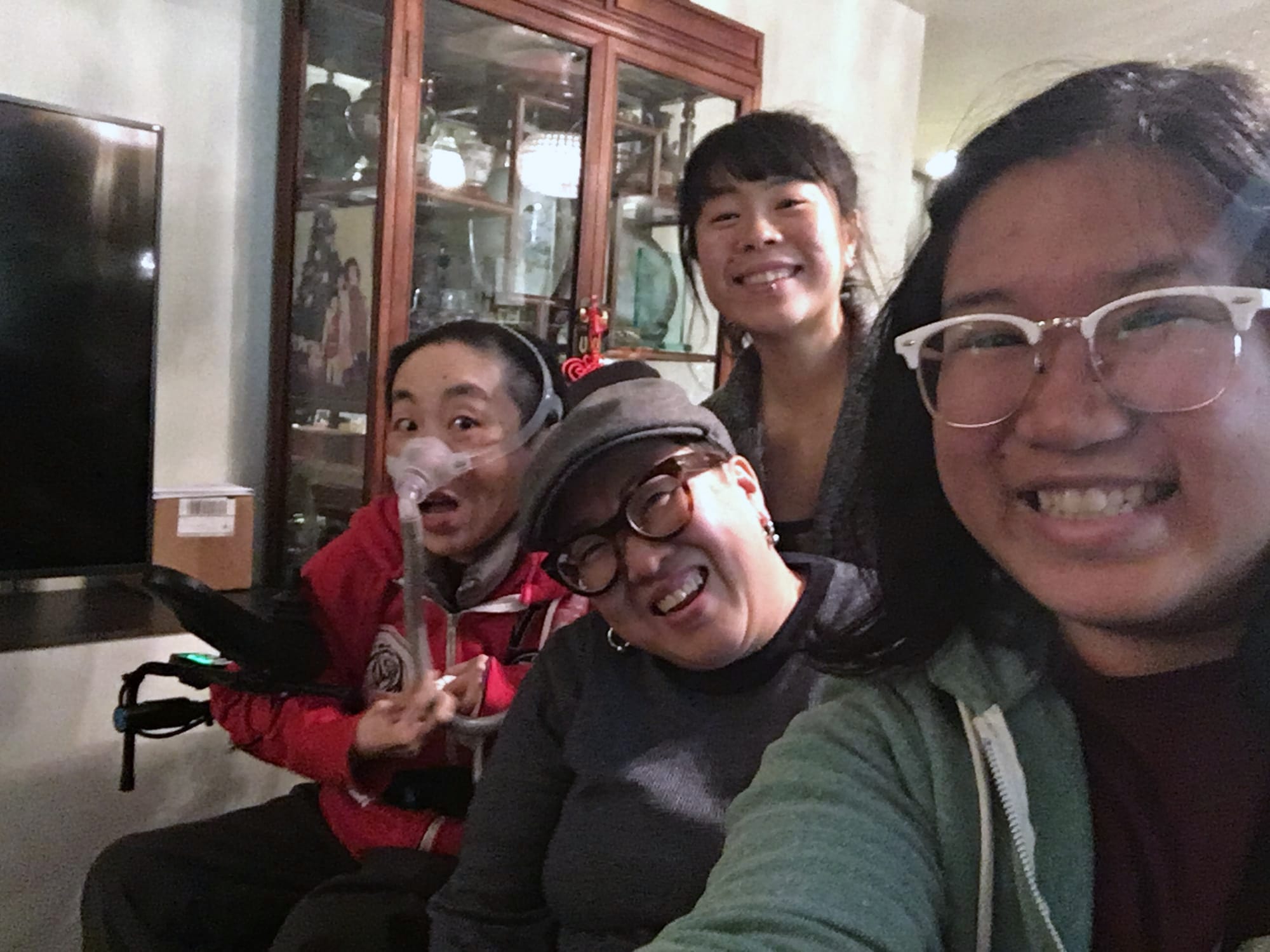 Four people smile at a camera, selfie-style. From left: a woman wearing a ventilator and a red hoodie; a woman in a newsie cap ad grey sweater; a woman with bangs wearing a grey blazer; and a person with black hair and white glasses.