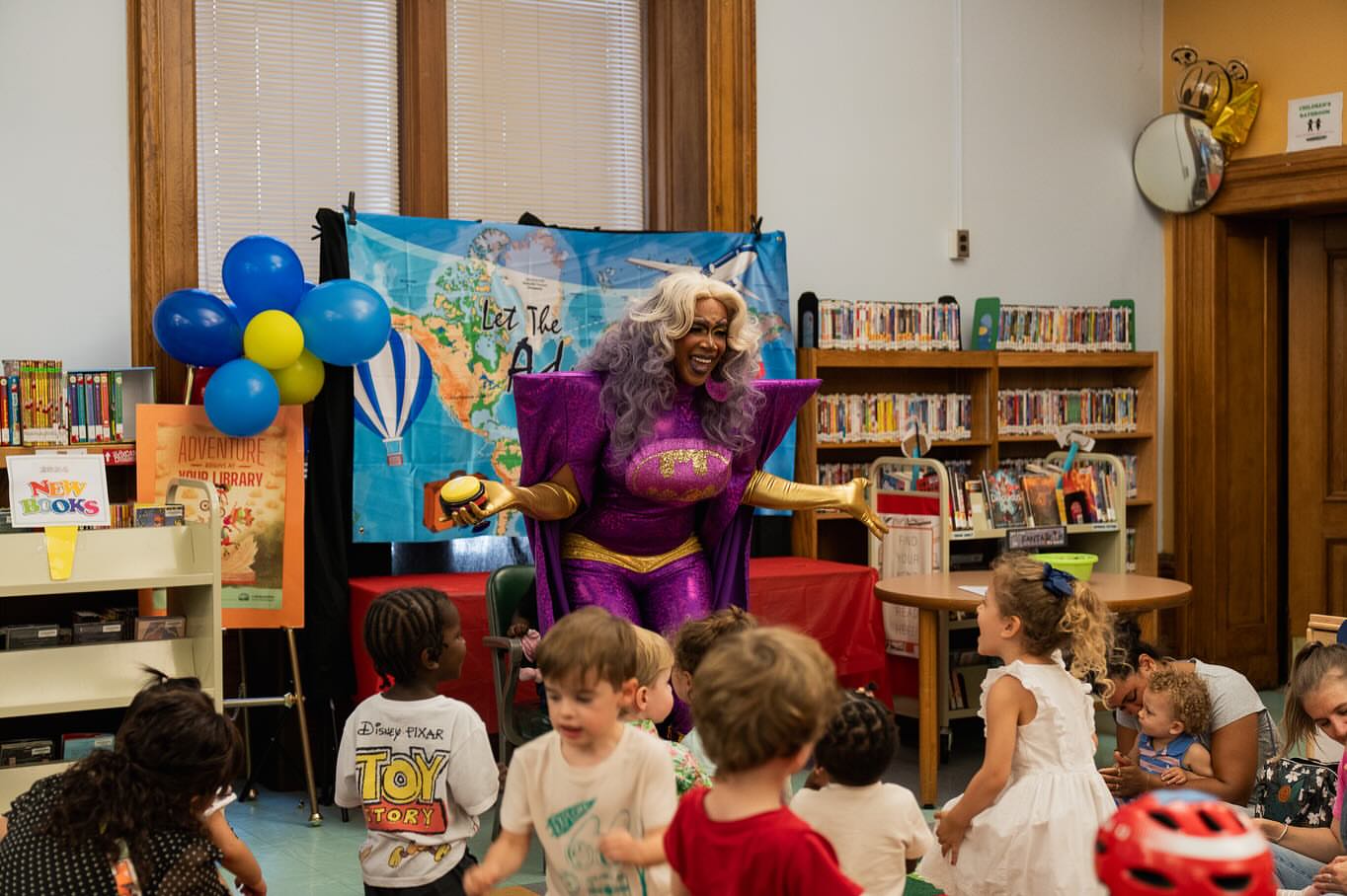 a beautiful Black drag queen in a purple outfit smiles while talking to young children in a library