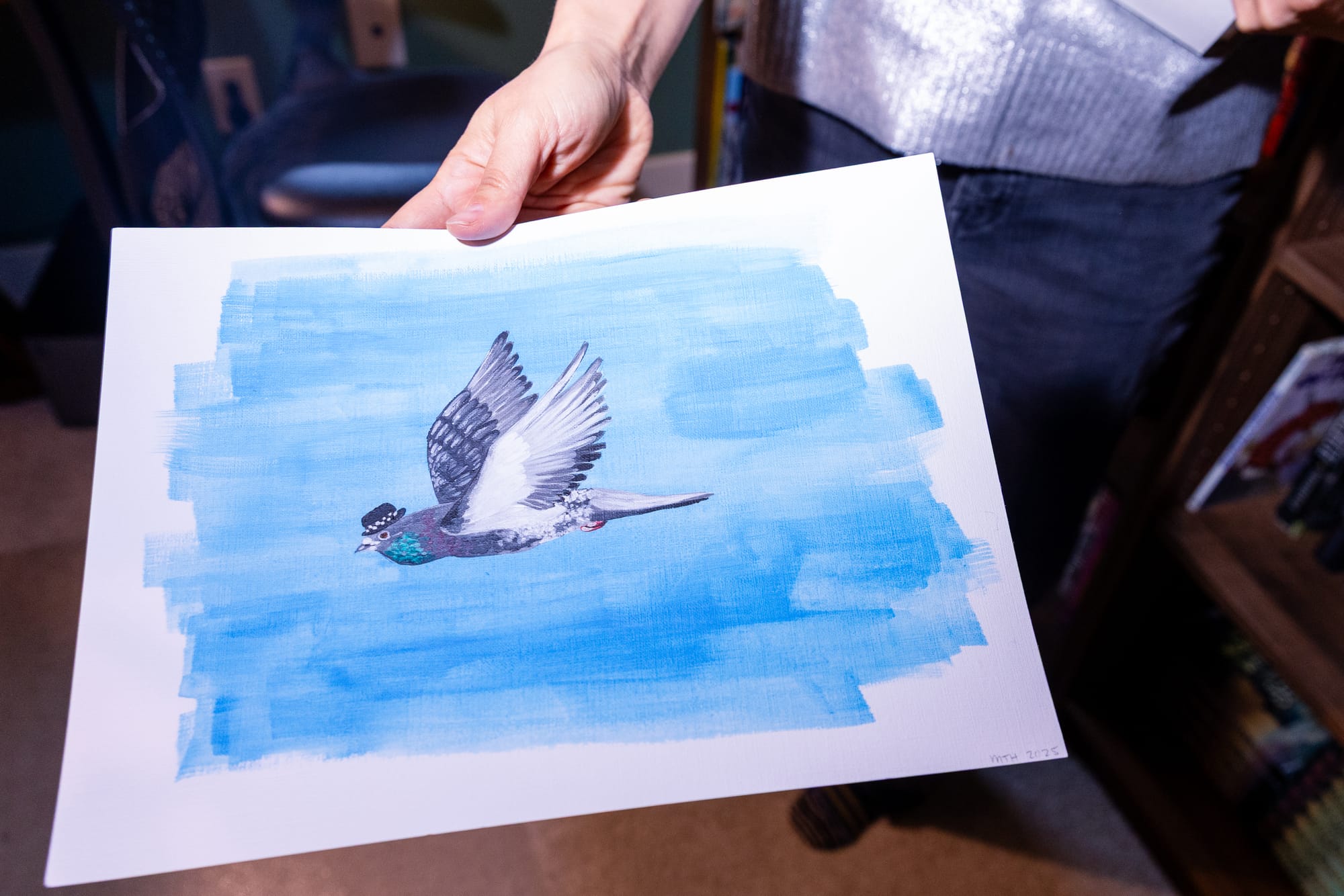 A hand holding a painting of a pigeon, mid flight. The pigeon is wearing a bowler hat.
