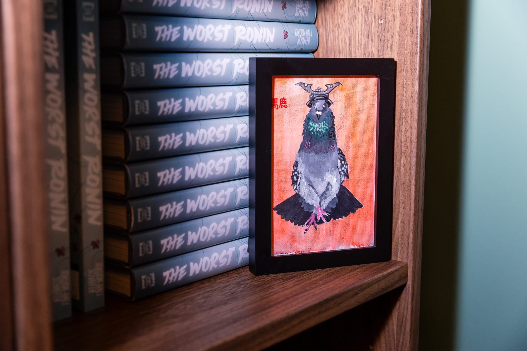 A painting of a pigeon with crossed legs, wearing a japanese warrior helmet. In the background, are a stack of copies of the book The Worst Ronin.