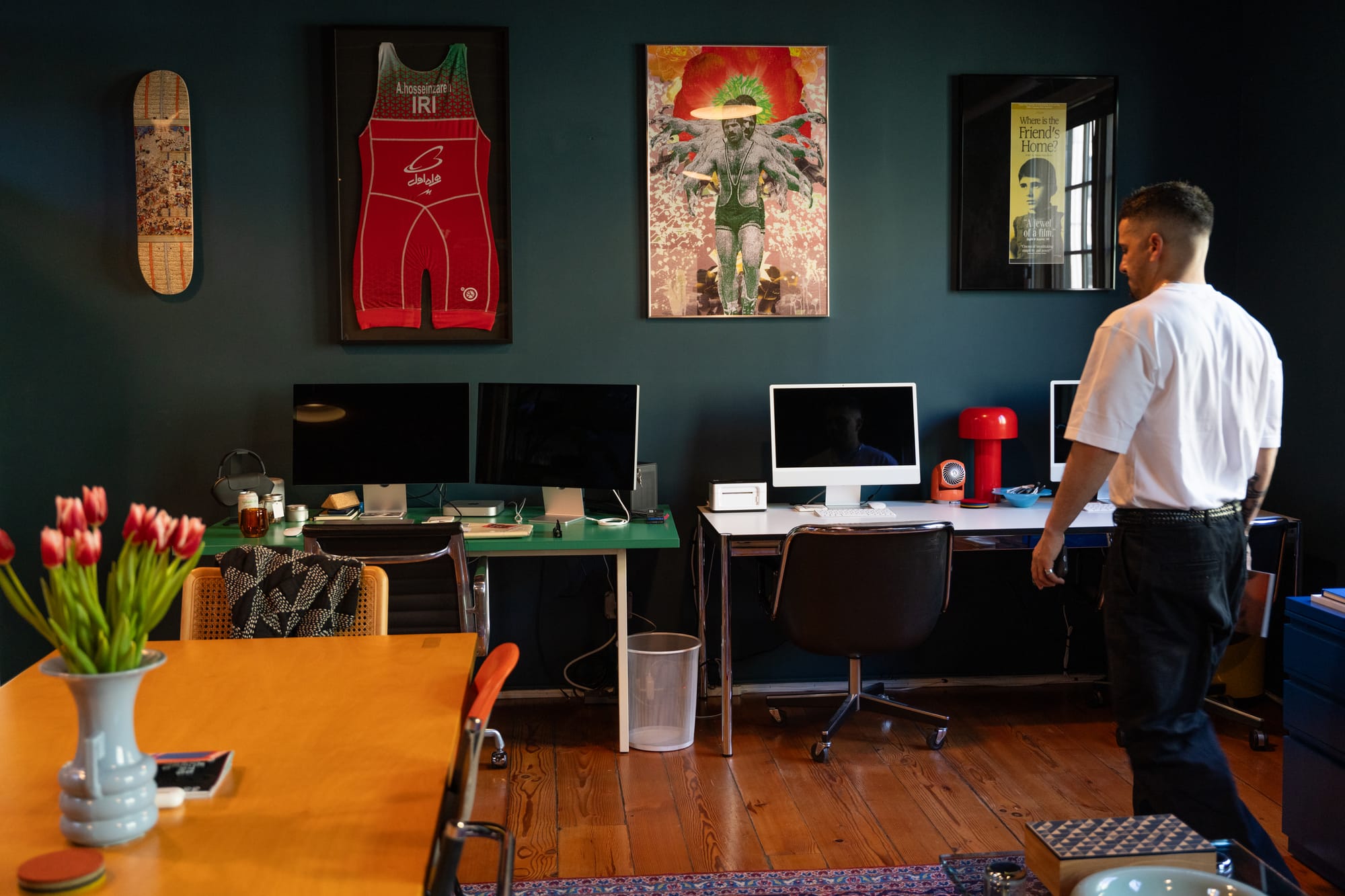 Man in a white t-shirt standing in front of computer desks. Hanging on the wall are a skateboard, Iranian wrestling jersey, poster, and film poster.
