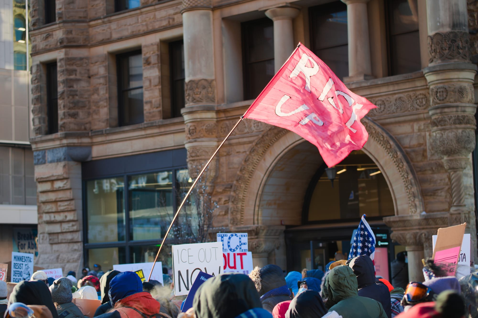 A protest with people holding signs that say 'ice out now' in. front of a brick building.