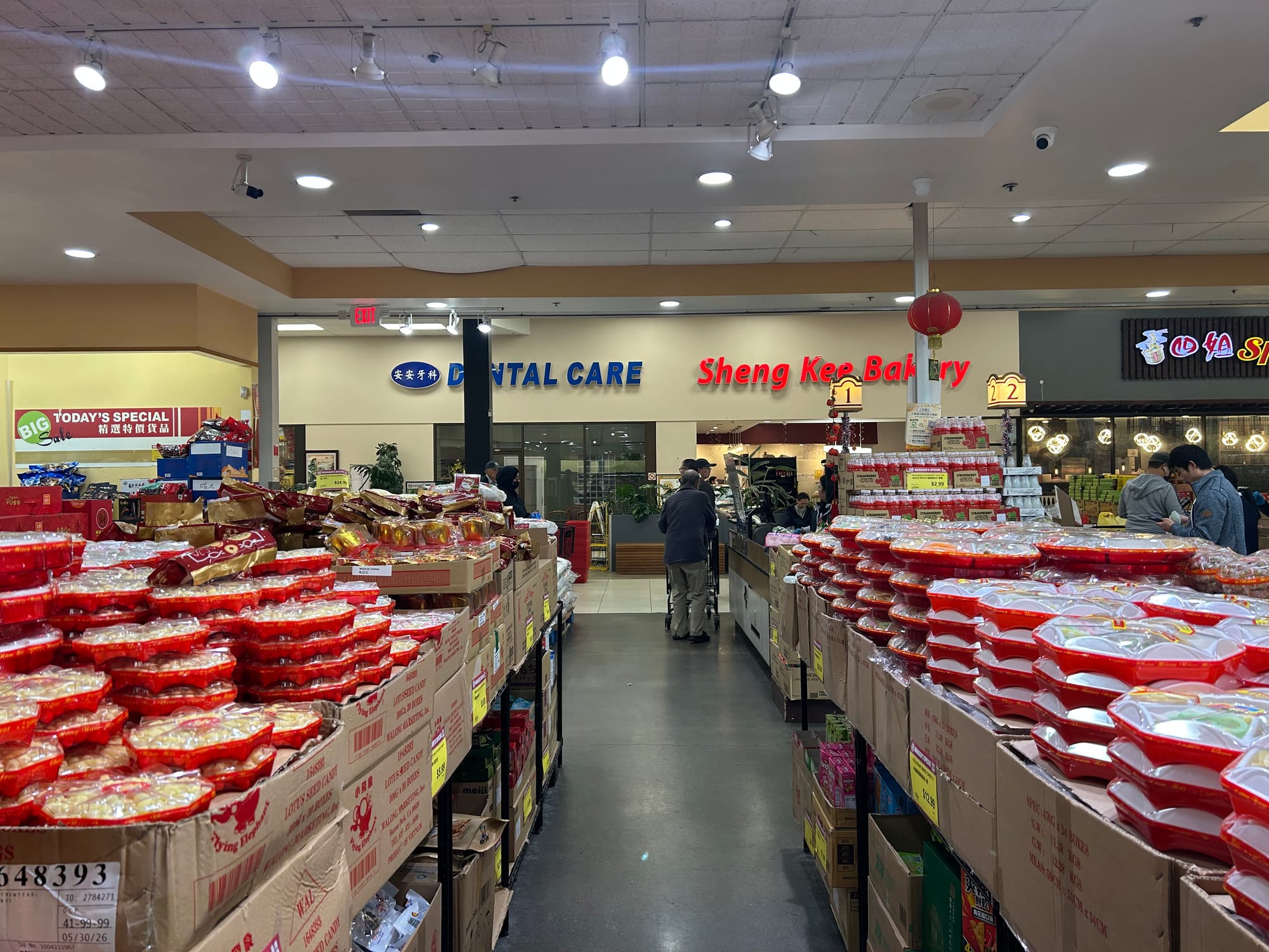 The entrance of Ranch 99, with shelves holding red trays of sweets and snacks. In the background are storefronts for a dental practice and a bakery.