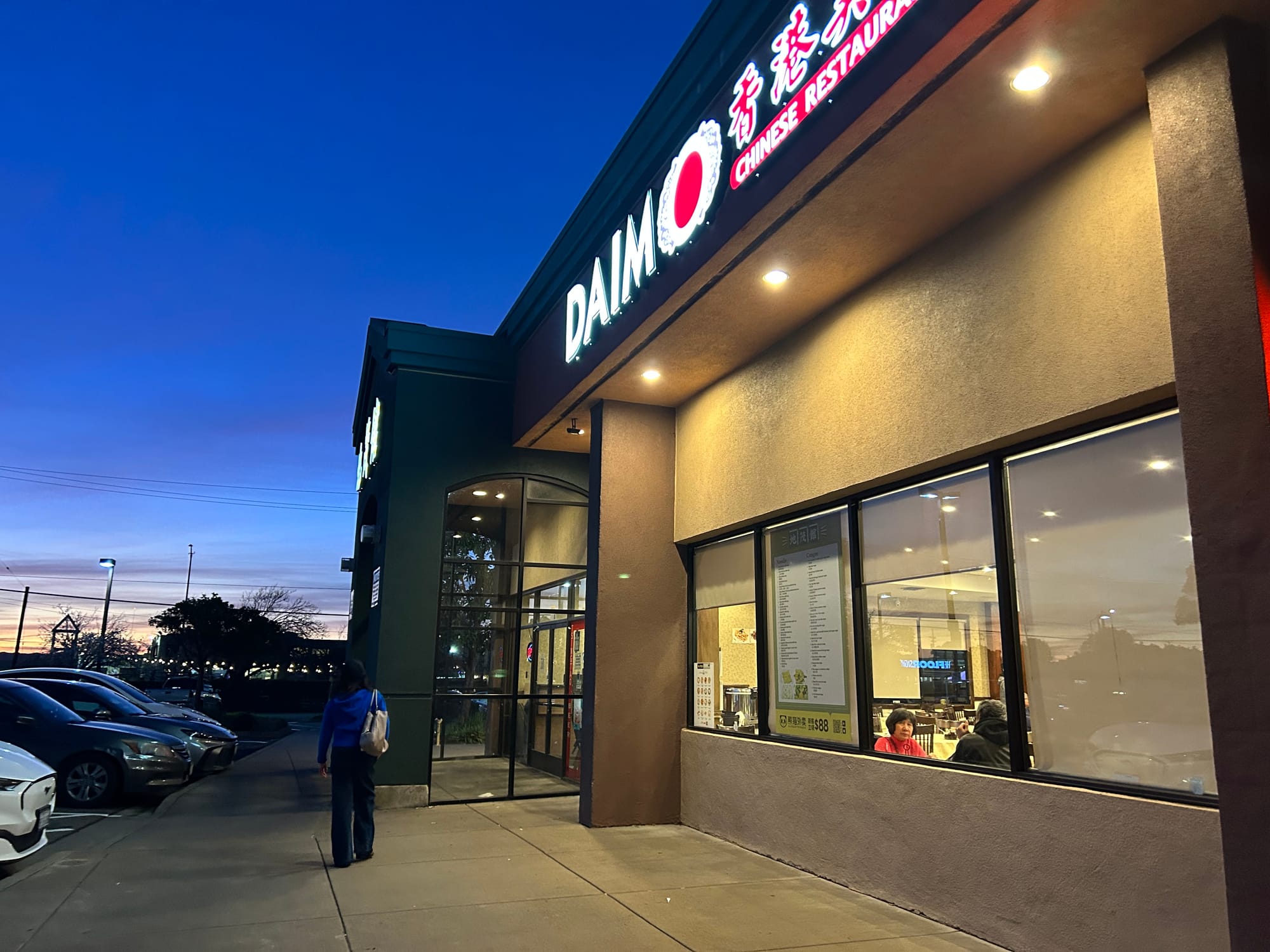 A woman in a blue sweater walks in front of a restaurant, Daimo, at dusk.