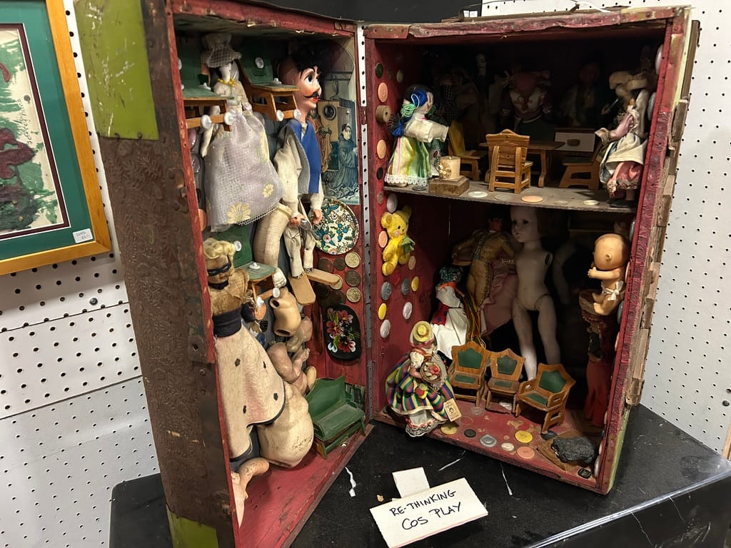 A trunk sits open on its side. Inside are dolls of various sizes, lots of small chairs, and buttons on the walls.