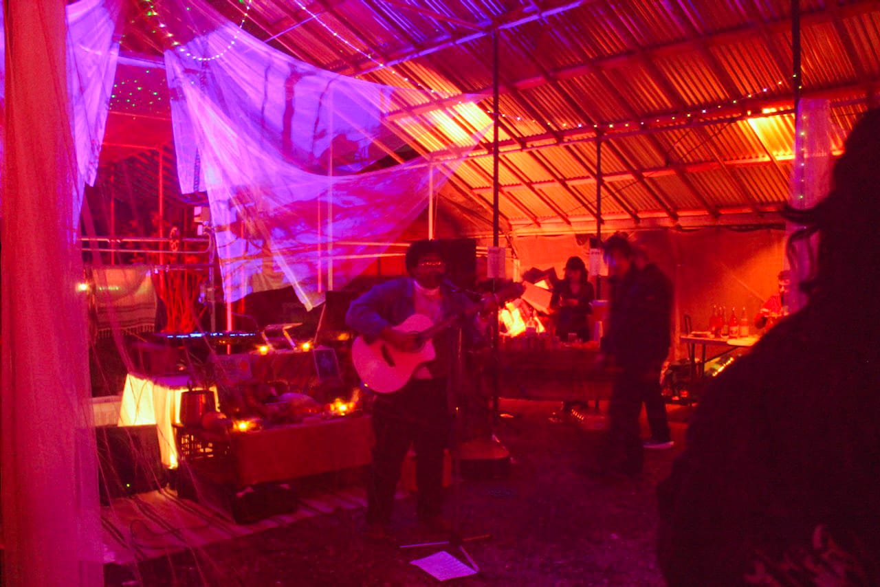 In a hazy, dark warehouse space a person holds an acoustic guitar and sings into a microphone.