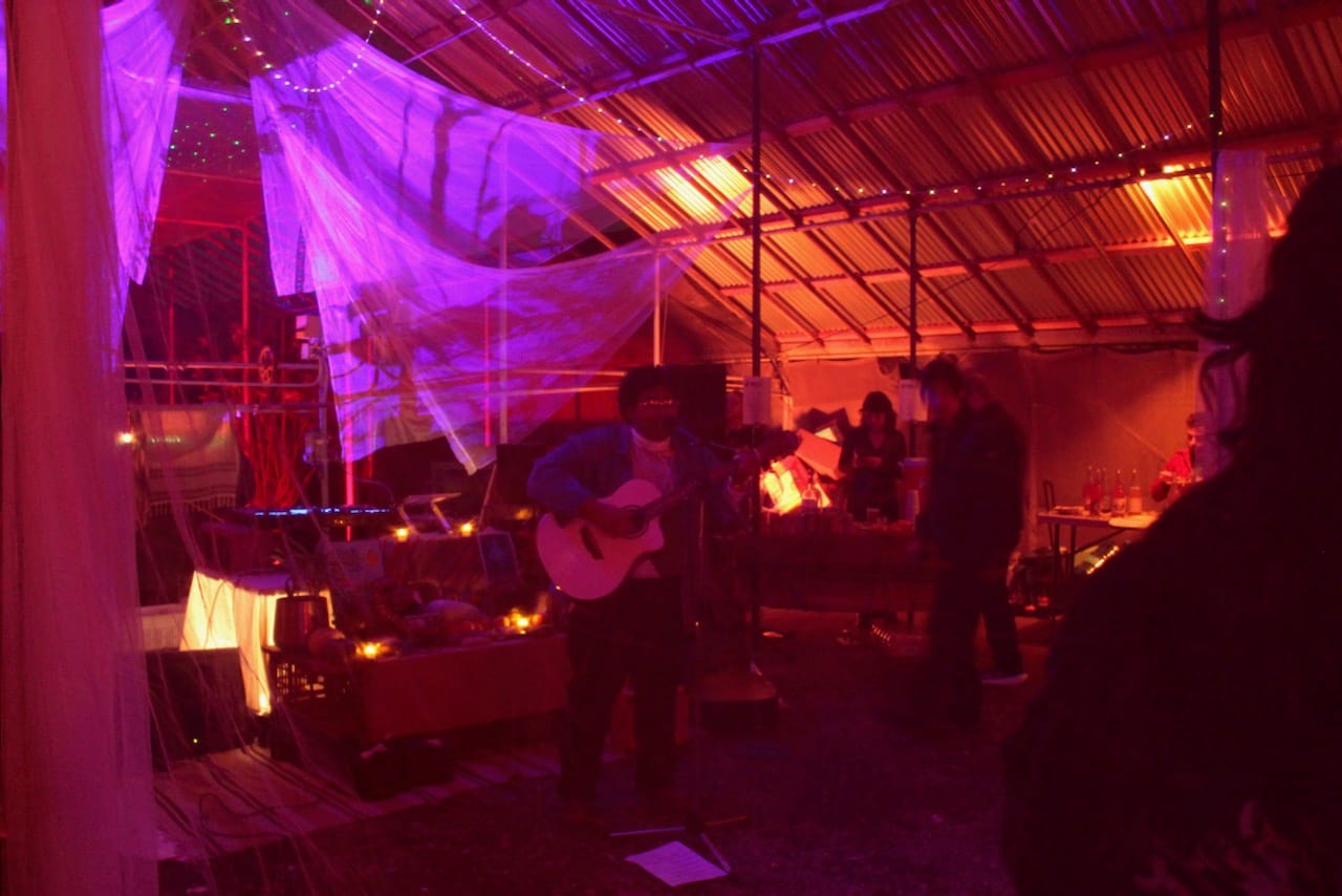 In a hazy, dark warehouse space a person holds an acoustic guitar and sings into a microphone.