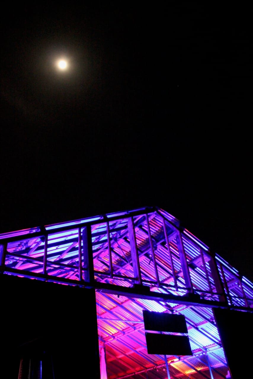 A warehouse space is lit by pink and orange lights, and the moon shines against the dark sky in the background.