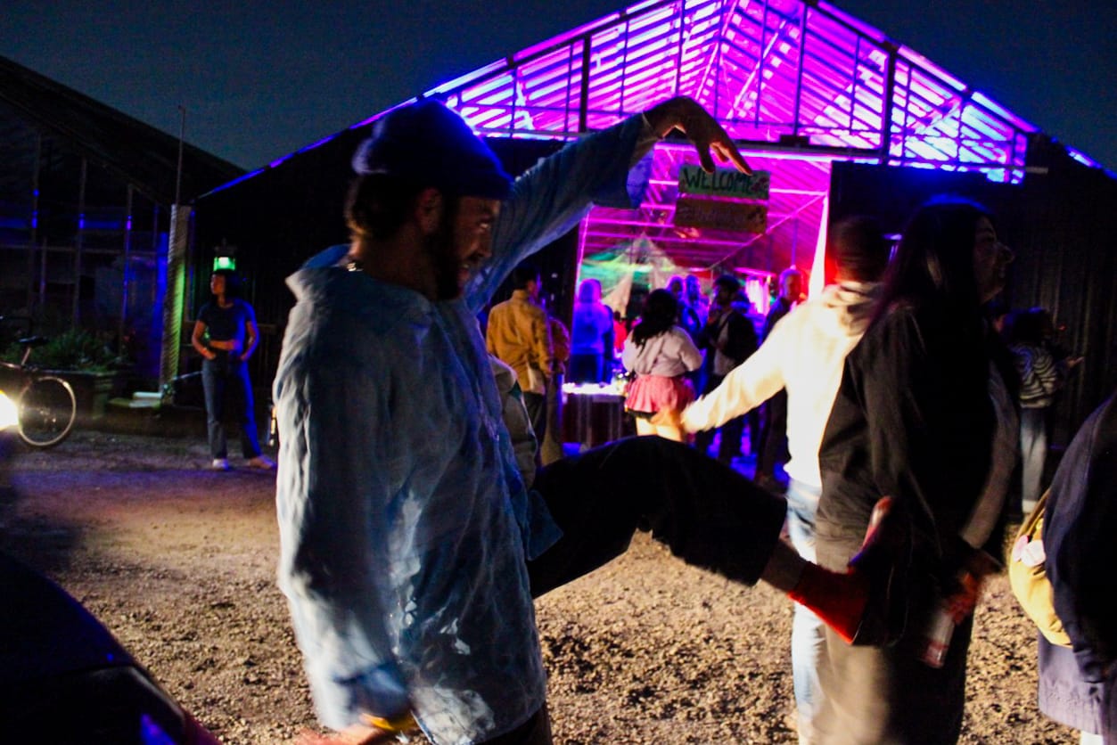 A person in the foreground points at a sign and raises his leg, while people mill around in the background.