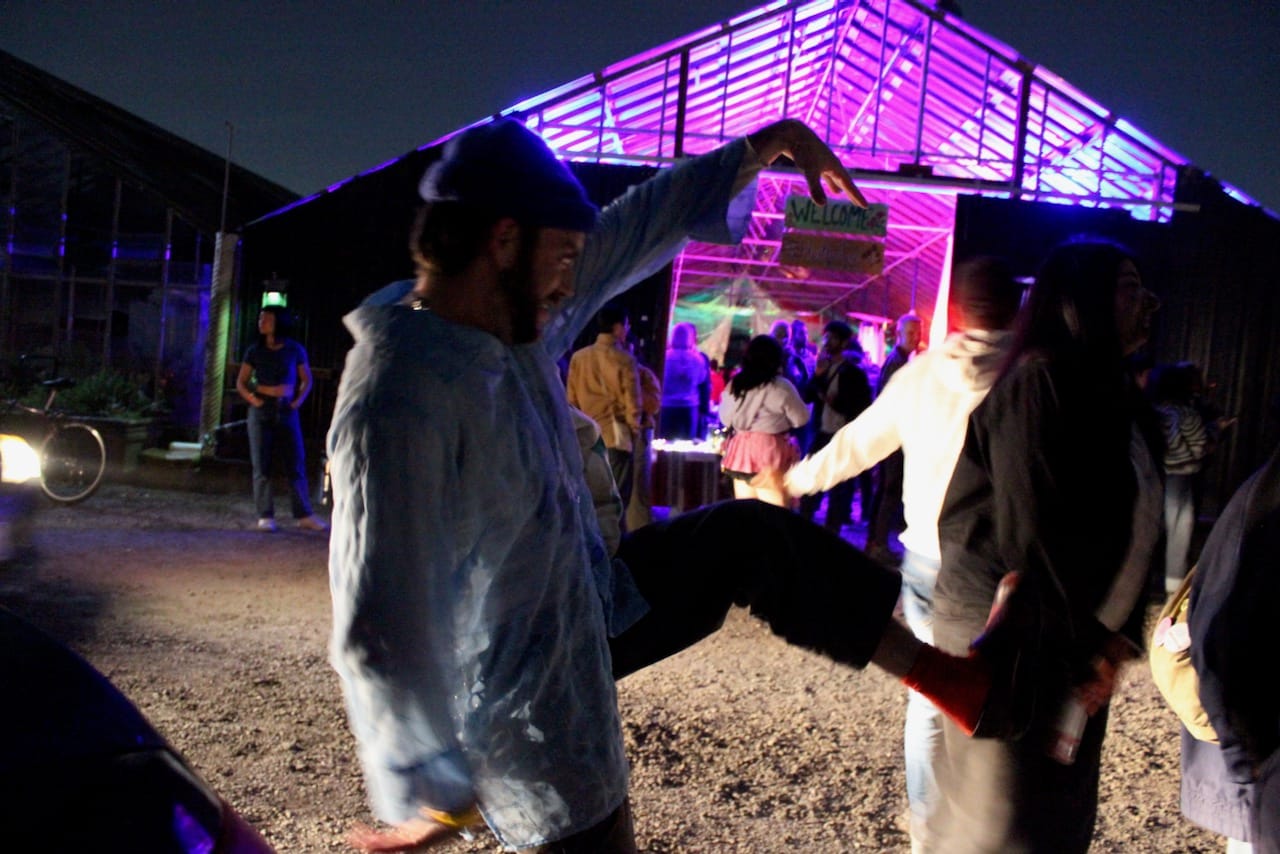A person in the foreground points at a sign and raises his leg, while people mill around in the background.