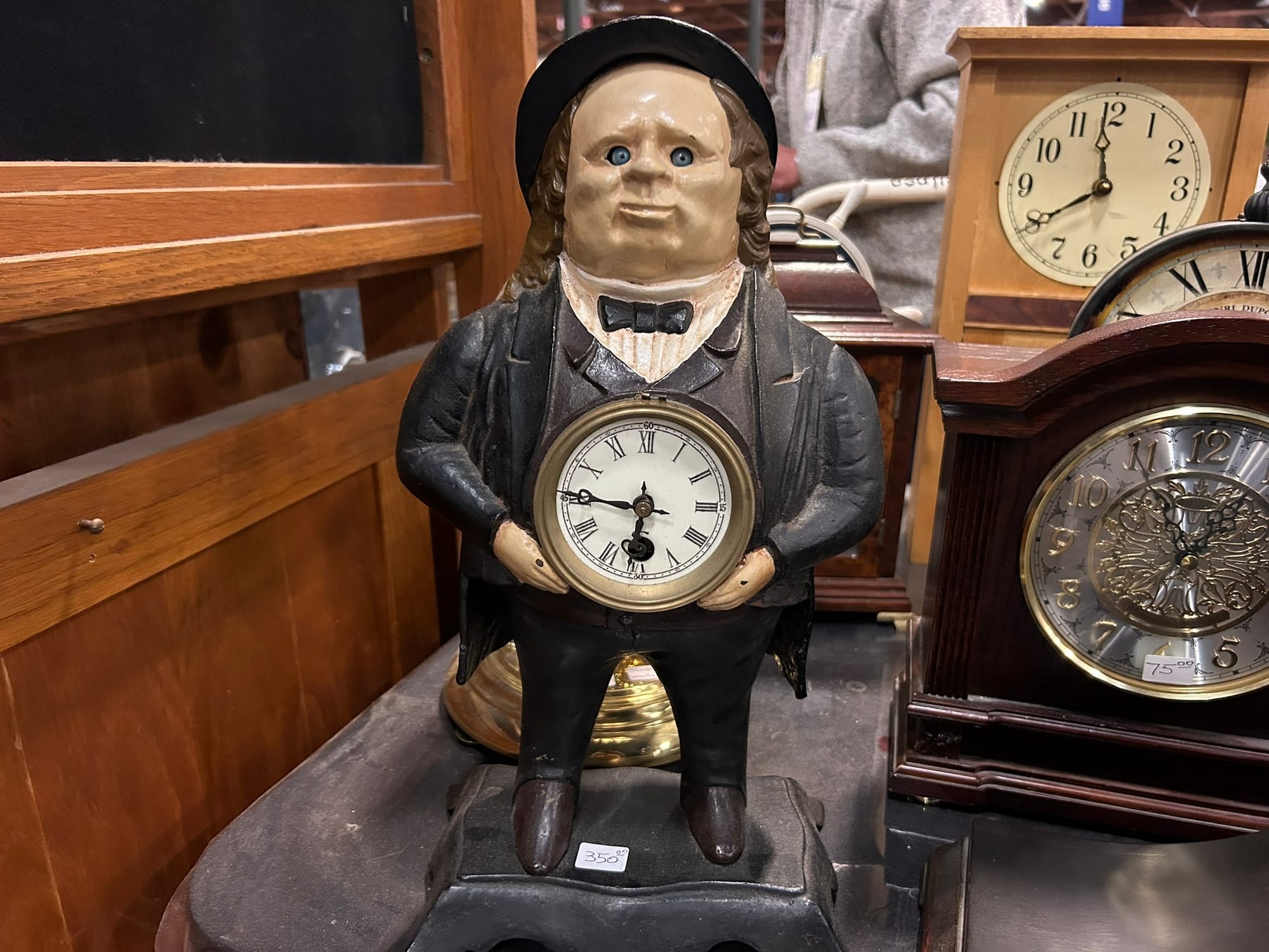 On a table filled with clocks, a statue of a man in a suit, with long brown hair and a hat, holds a small gold clock.