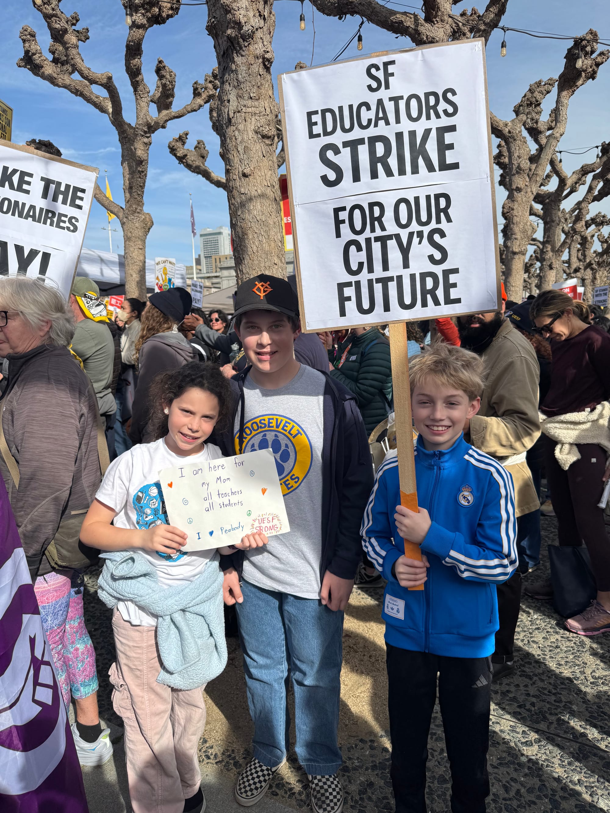 three kids at a protest, one holding a sign that says 'i am here for my mom all teachers all students i <3 peabody, uesf strong,' another is holding a sign that reads 'sf educators strike for our city's future'