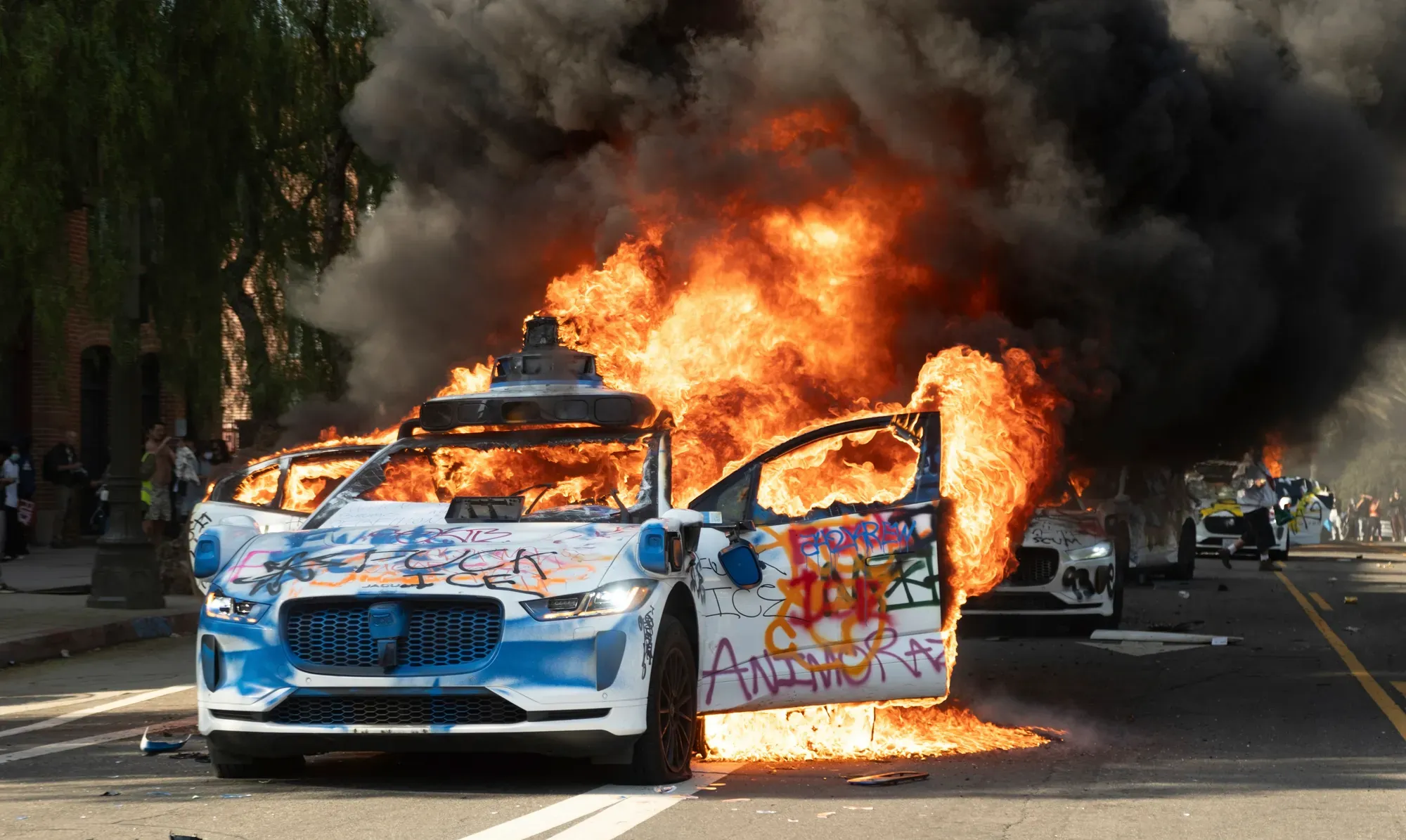 A Waymo vehicle is fully engulfed in flames on a road. Spraypaint is on the doors and body of the car, reading "Fuck Ice" on the hood