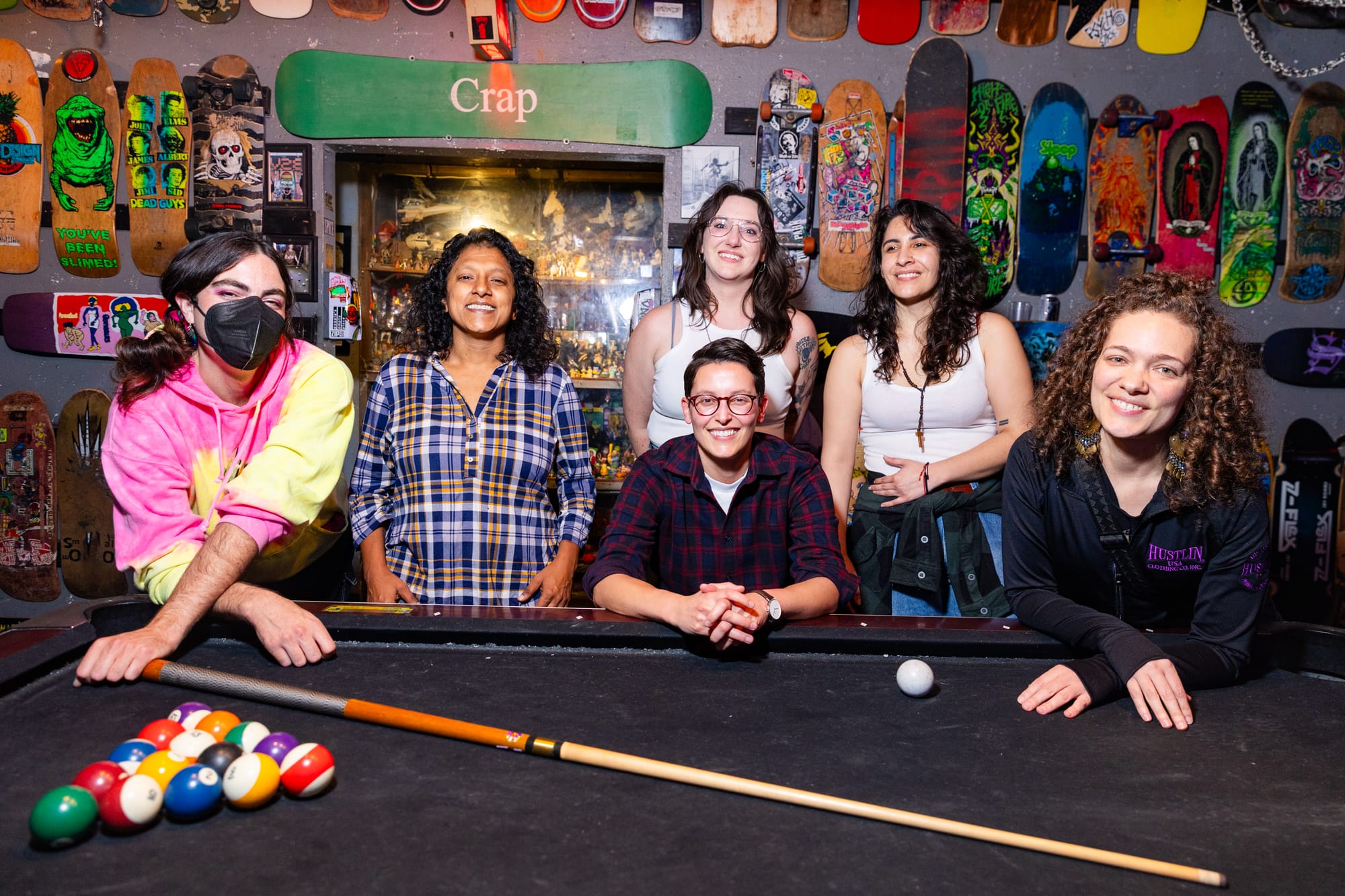 a group portrait of six people all smiling behind a pool table. They are the East Bay Baddie Billiards, a group working to make safer spaces for women, nonbinary, queer and trans folks who want to play pool