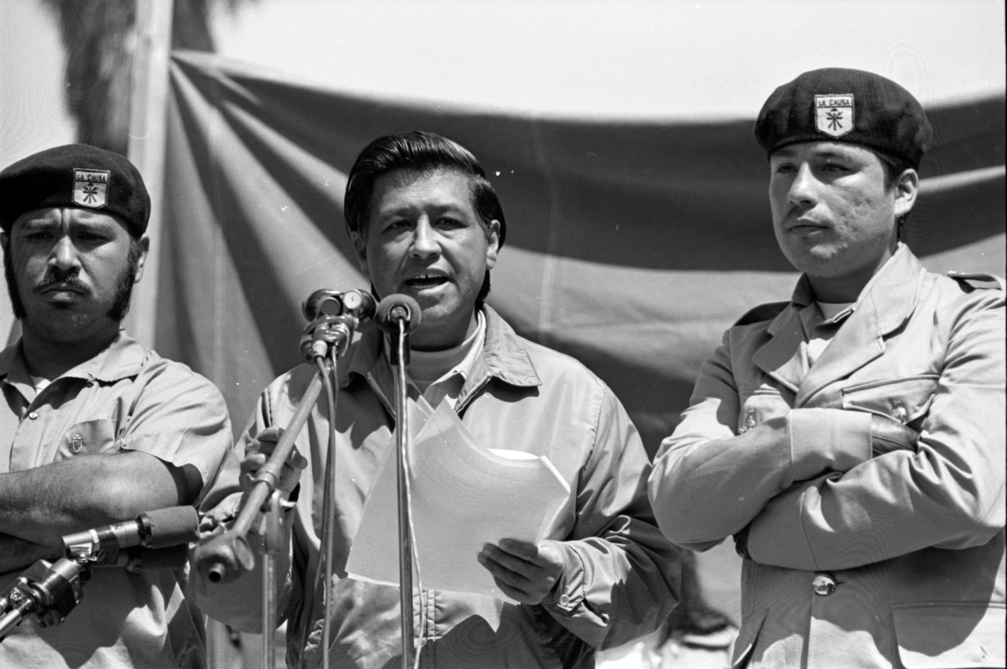 a black and white photo of Cesar Chavez, a Mexican American man, speaking into a microphone, with two men standing on either side of him in dark berets