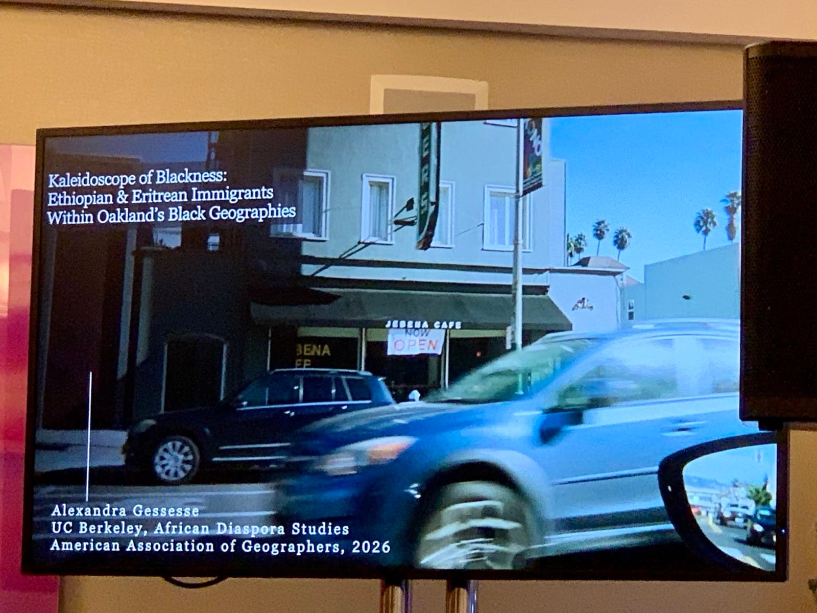 A screen that shows a car driving by and says āKaleidoscope of Blackness: Ethiopian and Eritrean Immigrants within Oakland's Black Geographies." Below that it says "Alexandra Gessesse UC Berkeley, African Diaspora Studies American Association of Geographers, 2026"