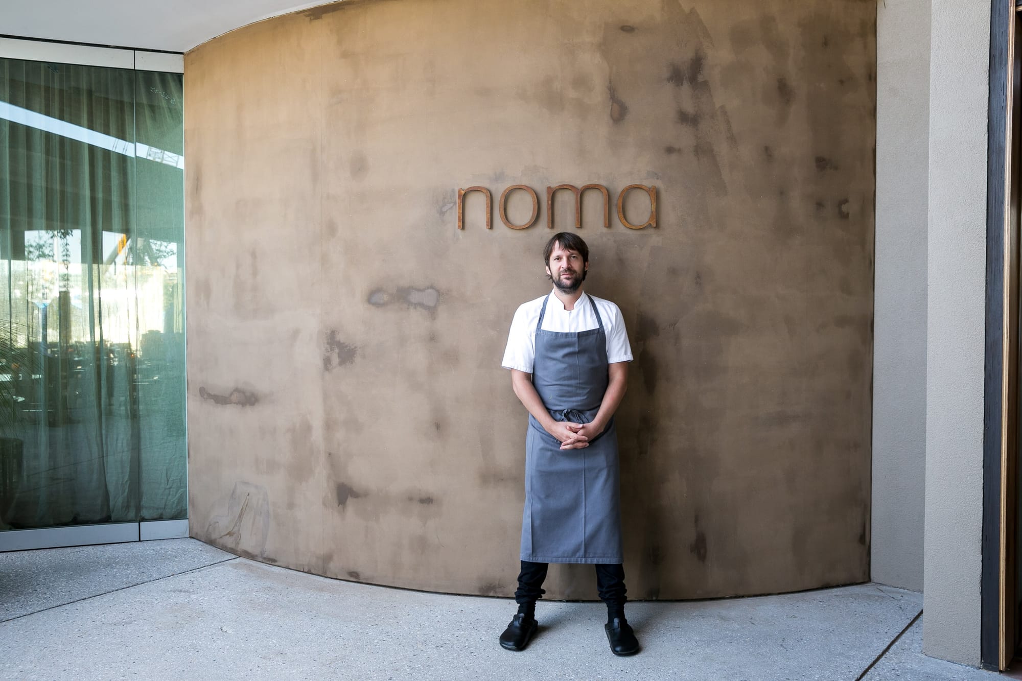 a white man in a grey chef's apron in front of a tan wall that reads 'noma'