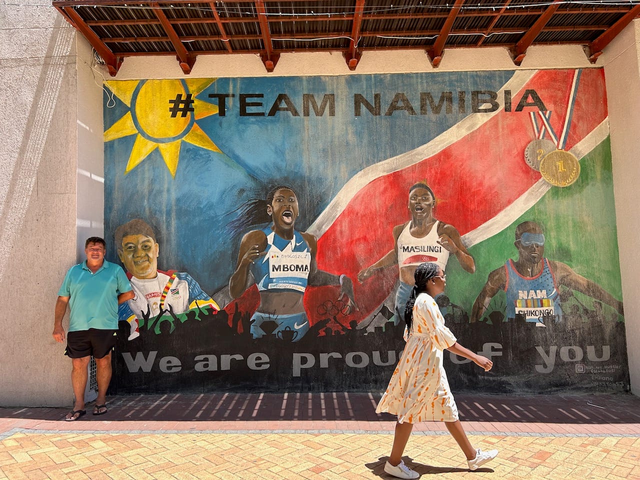 A man stands in front of a mural that depicts him next to three athletes. At the top the mural says "Team Namibia."