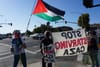 people are seen from behind standing on a street corner holding banners that read 'stop starving gaza' and waving a Palestinian flag