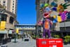A colorful statue of a bipedal creature with big ears and a huge mouth stands on a red plinth in front of an urban boulevard. A skybridge says "SJSU."