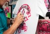 Everett Regua uses an airbrush on a white t-shirt to draw a black outline over a pink cross.