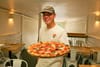 A smiling man in a baseball hat and white t-shirt holds a pepperoni and margherita pizza inside of a restaurant.
