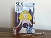 A book called "Sex Change and the City" on a table with a vial of testosterone cypionate and a used syringe.