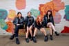 four women in black leather jackets and boots sit on a bench in front of a mural of colorful roses
